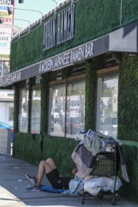 A homeless person sits on the sidewalk outside of Urban Ramen Bar at Sunset Blvd and Fuller Ave. in Los Angeles, Aug. 16, 2022. (Photo by Ringo Chiu)